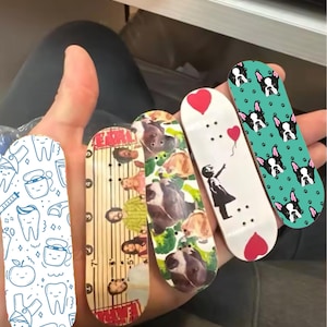 Pode incluir: Cinco mini skateboards com designs diferentes. Um tem um padrão de dentes azul e branco, outro tem uma régua com um padrão de linhas pretas e brancas, outro tem uma estampa de animais coloridos, outro tem um fundo branco com um coração vermelho e uma menina segurando um balão, e o último tem um fundo verde com um padrão de cachorro preto e branco.