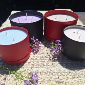 Puede incluir: Cuatro velas en recipientes de diferentes colores. Dos velas están en recipientes negros, una está en un recipiente rojo y una está en un recipiente morado. Las velas están todas encendidas y tienen tres mechas cada una.