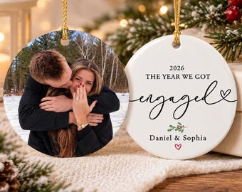 May include: Two round ceramic ornaments with gold hanging strings. One ornament features a photograph of a couple embracing in a snowy forest, with one person showing an engagement ring. The other white ornament displays the text 2026 THE YEAR WE GOT engaged, Daniel & Sophia, a small mistletoe graphic, and a red heart.