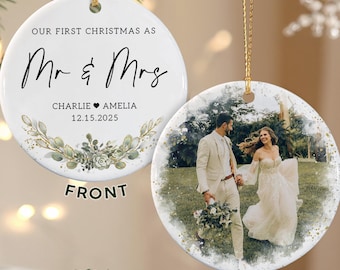 May include: A white ceramic Christmas ornament with gold string. The front reads "OUR FIRST CHRISTMAS AS Mr & Mrs" with names and a date. The back features a photo of a couple. The ornament is decorated with greenery.