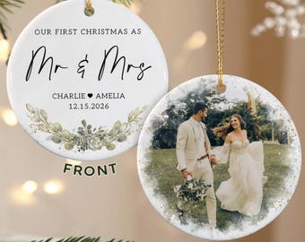 Pode incluir: Dois ornamentos redondos de cerâmica com cordões dourados. A frente apresenta o texto "OUR FIRST CHRISTMAS AS Mr & Mrs", os nomes "CHARLIE & AMELIA", a data "15.12.2026" e uma coroa botânica verde. O verso mostra uma foto de um casal.