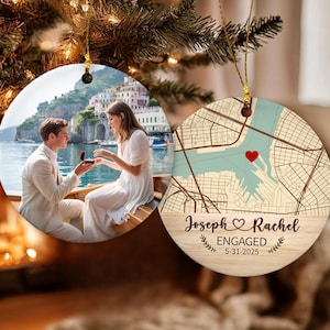 May include: Two round ceramic ornaments hanging from a Christmas tree. One features a photo of a couple, the other a map with a red heart and the text "Joseph & Rachel ENGAGED 5-31-2025".