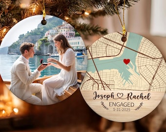 May include: Two round ceramic ornaments hanging on a tree. One shows a couple on a boat with a coastal town background. The other features a map with a red heart, the text Joseph & Rachel, ENGAGED, and the date 5-31-2025 on a wood grain base.