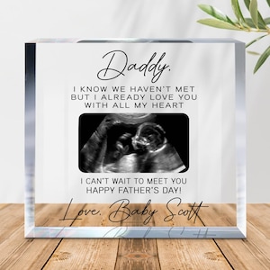 May include: A clear acrylic block with the text "Daddy" at the top. Below, it reads "I KNOW WE HAVEN'T MET BUT I ALREADY LOVE YOU WITH ALL MY HEART." An ultrasound image is centered, with "I CAN'T WAIT TO MEET YOU HAPPY FATHER'S DAY!" below. The bottom reads "Love. Baby Scott."