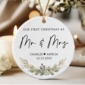May include: A round white ceramic ornament featuring the black text OUR FIRST CHRISTMAS AS Mr & Mrs, the names CHARLIE and AMELIA separated by a heart, and the date 12.15.2025. A green botanical wreath with gold accents sits below.