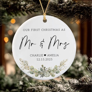 May include: A round white ceramic ornament featuring the black text "OUR FIRST CHRISTMAS AS Mr & Mrs" with the names "CHARLIE &hearts; AMELIA" and the date "12.15.2025" printed below. A gold string is attached to the top, and a green botanical illustration.