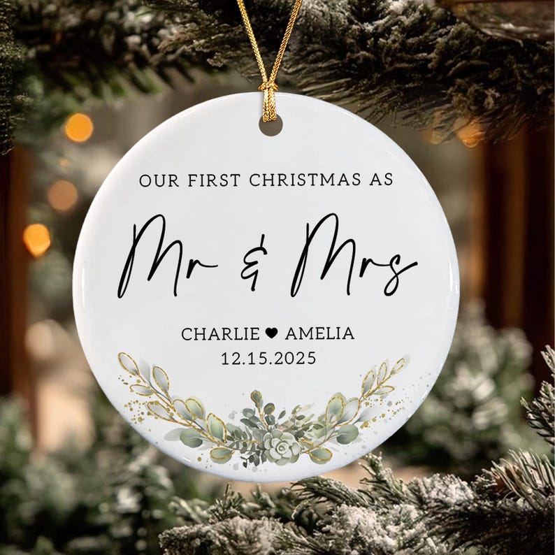 May include: A round white ceramic ornament featuring the black text "OUR FIRST CHRISTMAS AS Mr & Mrs" with the names "CHARLIE &hearts; AMELIA" and the date "12.15.2025" printed below. A gold string is attached to the top, and a green botanical illustration.