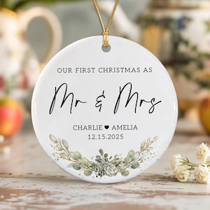 May include: A round white ceramic Christmas ornament featuring the black text OUR FIRST CHRISTMAS AS Mr & Mrs, the names CHARLIE and AMELIA separated by a heart, and the date 12.15.2025, accented with a green botanical wreath at the bottom.