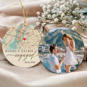 May include: A round ceramic ornament featuring a map of New York with a red heart, the names Daniel & Lauren, the word engaged, and the date 05.16.2026. A second round ornament displays a photograph of a proposal in a scenic outdoor setting.