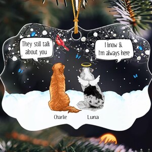 May include: A decorative ornament featuring a golden retriever named Charlie and a spotted dog named Luna with angel wings and a halo. The dogs sit in snow under a dark sky with a red cardinal and blue butterflies. Text reads: They still talk about you and I know & I'm always here.