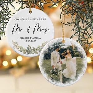 May include: Two round white ceramic ornaments with gold hanging strings. One ornament features the black text "OUR FIRST CHRISTMAS AS Mr & Mrs" with the names "CHARLIE & AMELIA" and date "12.15.2025" above a green watercolor botanical illustration.
