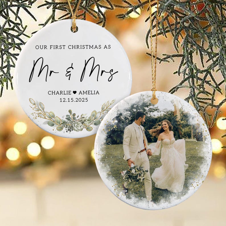 May include: Two round white ceramic ornaments with gold hanging strings. One ornament features the black text "OUR FIRST CHRISTMAS AS Mr & Mrs" with the names "CHARLIE & AMELIA" and date "12.15.2025" above a green watercolor botanical illustration.