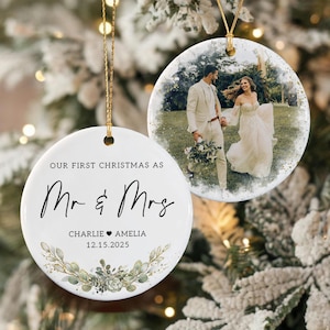 May include: Two round white ceramic ornaments with gold hanging strings. One features a printed wedding photograph of a couple, while the other displays the text "OUR FIRST CHRISTMAS AS Mr & Mrs", the names "CHARLIE &hearts; AMELIA", the date "12.15.2025", and a green botanical illustration.