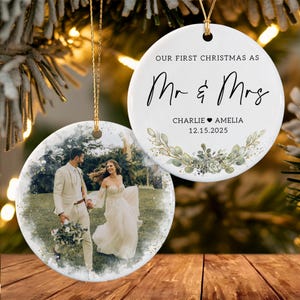 May include: Two round white ceramic ornaments with gold hanging strings. One ornament features a printed photo of a couple in wedding attire. The other displays the text "OUR FIRST CHRISTMAS AS Mr & Mrs", "CHARLIE &hearts; AMELIA", "12.15.2025", and a green botanical wreath.