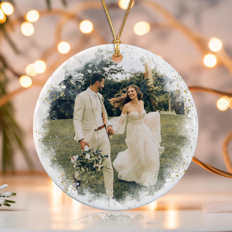 May include: A round white ceramic ornament featuring a printed photograph of a couple in wedding attire walking through a grassy field. The ornament has a gold string attached at the top for hanging and a watercolor-style border with gold speckles.