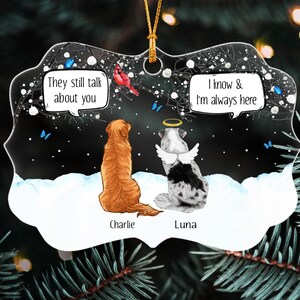May include: A decorative ornament featuring a golden dog named Charlie and a spotted dog named Luna with angel wings and a halo. The scene shows them in snow with a red cardinal and blue butterflies. Text reads: They still talk about you and I know & I'm always here.