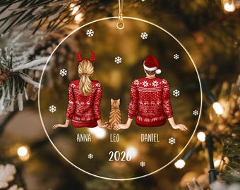 Puede incluir: Un adorno navideño redondo y transparente con una ilustración impresa de dos personas y un gato vistos desde atrás. Llevan jerséis rojos a juego con motivos. Los nombres Anna, Leo y Daniel están impresos debajo, con el año 2026 al pie.