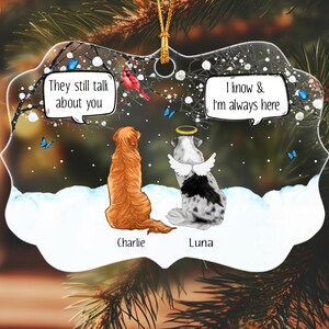 May include: A clear, scalloped-edge ornament featuring a golden retriever and a spotted dog with angel wings and a halo. The dogs sit in snow under a tree with blue butterflies and a red bird. Text bubbles read: They still talk about you and I know & I'm always here. Names Charlie and Luna are printed below.