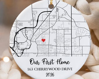 May include: A round white ceramic ornament featuring a black line map graphic with a small red heart icon. The text reads Our First Home, 163 CHERRYWOOD DRIVE, and 2026. A gold string is attached to the top for hanging the decorative piece.