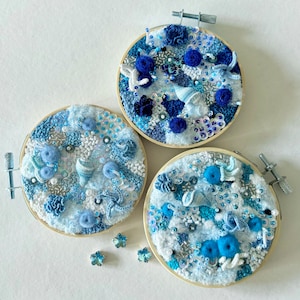 May include: Three round embroidery hoops featuring blue and white crocheted designs. The designs incorporate seashells, beads, and other ocean-inspired elements.
