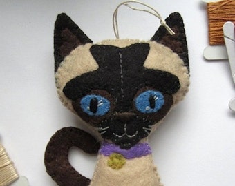 May include: A felt Siamese cat ornament with blue eyes, a purple collar with a gold charm, and a brown tail. The cat is sitting upright with its legs together. The ornament has a loop for hanging. The text "www.BayColonyDesigns.etsy.com" is printed on the bottom.