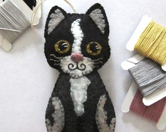 May include: A black and white felt cat ornament with yellow eyes and a pink nose. The cat is sitting with its paws tucked under its body. The ornament is hanging from a string. There are spools of thread in the background.