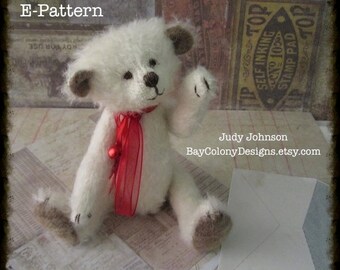 May include: White teddy bear with brown ears and a red ribbon around its neck. The bear is sitting on a piece of paper with a white envelope next to it. The text "E-Pattern" is visible in the top left corner of the image.