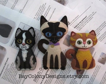May include: Three felt cat toys, a black and white cat, a brown and white cat, and a Siamese cat. The cats are all wearing collars and have embroidered faces. The text "Take a stake in my pattern?" is visible in the image.