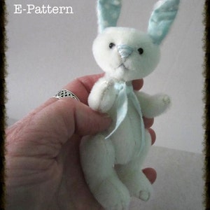 Op de afbeelding: Een wit pluche konijn met blauwe en witte stippenoren en een blauw lint om zijn nek. Het konijn wordt vastgehouden door een hand. De tekst "E-Pattern" staat in de linkerbovenhoek van de afbeelding. De tekst "Judy Johnson BayColonyDesigns.etsy.com" staat in het midden onderaan de afbeelding.