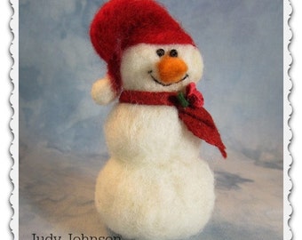 May include: A white felt snowman wearing a red hat and scarf with a red flower. The snowman is standing on a blue background. #9514 Judy Johnson BayColonyDesigns.com