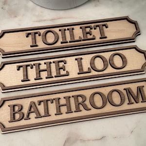 May include: Three wooden signs with dark brown lettering and trim. The signs read "TOILET", "THE LOO", and "BATHROOM". The signs are light brown with a dark brown border and lettering. The signs are rectangular with decorative edges.