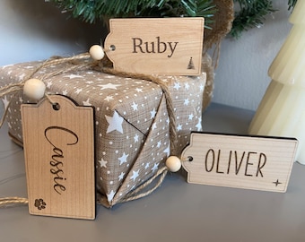 May include: Wooden gift tags with names "Ruby", "Cassie", and "Oliver" are displayed. The tags are attached to a wrapped gift with a star pattern. The tags have a wooden bead and twine. A small Christmas tree is in the background.