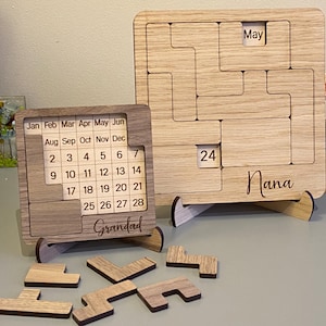 May include: Two wooden perpetual calendars with puzzle-piece designs. One shows May and the date 24, with "Nana" engraved below. The other shows a full month calendar with "Grandad" engraved below. Several puzzle pieces are scattered.