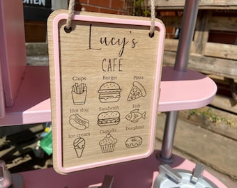May include: Wooden sign for a child's cafe, with the name "Lucy's Cafe" at the top. The sign features illustrations of food items like chips, burgers, pizza, hot dogs, sandwiches, fish, ice cream, cupcakes, and doughnuts. The sign has a pink border.