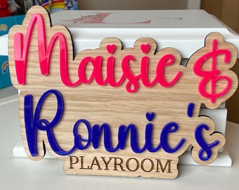 May include: A wooden sign with the words "Maisie & Ronnie's Playroom" in pink and blue lettering. The sign is decorated with small hearts.
