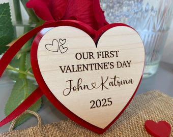 May include: A red heart-shaped wooden ornament with the text "Our First Valentine's Day John & Katrina 2025" engraved on it. The ornament is decorated with a red ribbon and two small hearts.
