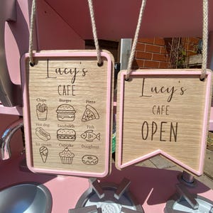 May include: Wooden cafe signs with pink trim and rope hangers. One sign says "Lucy's Cafe" and lists food items like chips, burgers, and pizza. The other sign says "Lucy's Cafe OPEN". The signs are part of a pink toy kitchen.