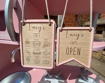 May include: Wooden cafe signs with pink trim and rope hangers. One sign says "Lucy's Cafe" and lists food items like chips, burgers, and pizza. The other sign says "Lucy's Cafe OPEN". The signs are part of a pink toy kitchen.
