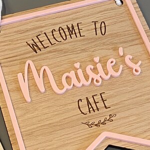 Puede incluir: Letrero de madera con borde rosa, que dice "Bienvenido a Maisie's Cafe" con un peque&ntilde;o dise&ntilde;o de hojas en la parte inferior.