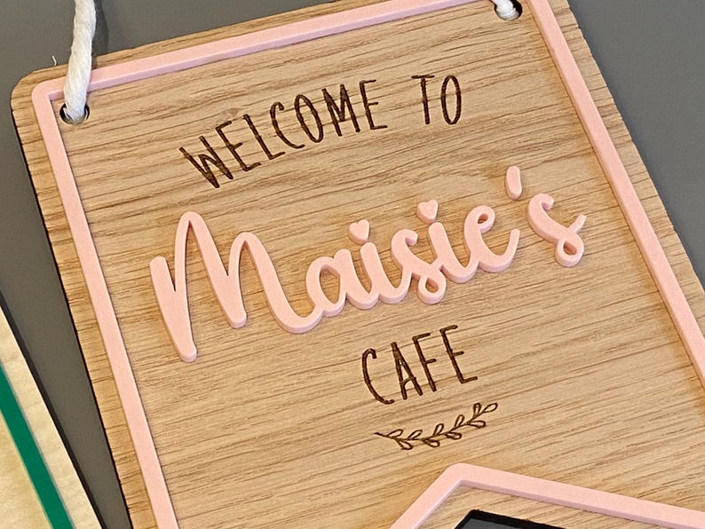 Puede incluir: Letrero de madera con borde rosa, que dice "Bienvenido a Maisie's Cafe" con un peque&ntilde;o dise&ntilde;o de hojas en la parte inferior.