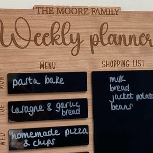 May include: A wooden weekly planner with black chalkboard sections for writing a menu and shopping list. The text "THE MOORE FAMILY" and "Weekly planner" is engraved on the wood. The chalkboard sections have handwritten text for the week's meals and grocery items.