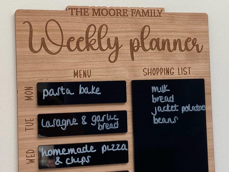 May include: A wooden weekly planner with black chalkboard sections for writing a menu and shopping list. The text "THE MOORE FAMILY" and "Weekly planner" is engraved on the wood. The chalkboard sections have handwritten text for the week's meals and grocery items.