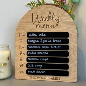 May include: A wooden weekly menu board with a rounded arch top. The board has a light wood finish and features a black chalkboard with the days of the week and a meal plan written in white chalk. The text "Weekly Menu" is engraved on the board. The board is labelled "The Moore Family".