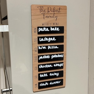 May include: Wooden weekly menu board with the text "The Robert Family" and "WEEKLY MENU". The menu includes daily meal options like pasta bake, lasagna, and roast dinner. The board is attached to a refrigerator.