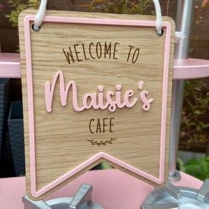Puede incluir: Un letrero de madera marr&oacute;n y rosa con un borde rosa y el texto "Bienvenido a Maisie's Cafe" con un peque&ntilde;o dise&ntilde;o de hojas.