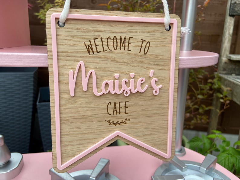 Puede incluir: Un letrero de madera marr&oacute;n y rosa con un borde rosa y el texto "Bienvenido a Maisie's Cafe" con un peque&ntilde;o dise&ntilde;o de hojas.