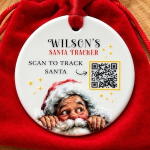 May include: A round white ceramic ornament featuring the text WILSON'S SANTA TRACKER and SCAN TO TRACK SANTA with a QR code, a graphic of Santa Claus peeking over the bottom edge, and yellow stars, suspended by a red ribbon over a red pouch.