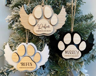 May include: Three wooden paw print ornaments with angel wings. The ornaments are painted white, black, and natural wood. The ornaments are personalized with the names "Rufus" and "Murrey" and the phrase "Forever in our hearts".
