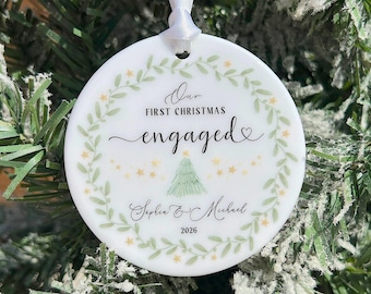 May include: A round white ceramic ornament featuring a green leaf wreath, small yellow stars, and a green Christmas tree illustration. The text reads Our FIRST CHRISTMAS engaged, Sophia & Michael, and 2026. A white ribbon is attached to the top.
