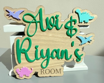 May include: A wooden wall sign with the green script text Avi & Riyan's ROOM. The sign features light purple, teal, light blue, and pink dinosaur and pterodactyl cutouts attached to the natural wood surface with a light wood grain finish.